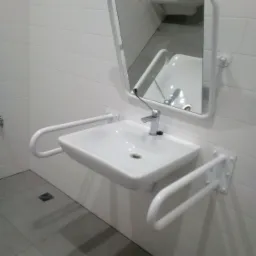 Biała umywalka dla niepełnosprawnych z poręczami i lustrem, zamontowana w łazience w Łodzi, widoczna na tle białych płytek.