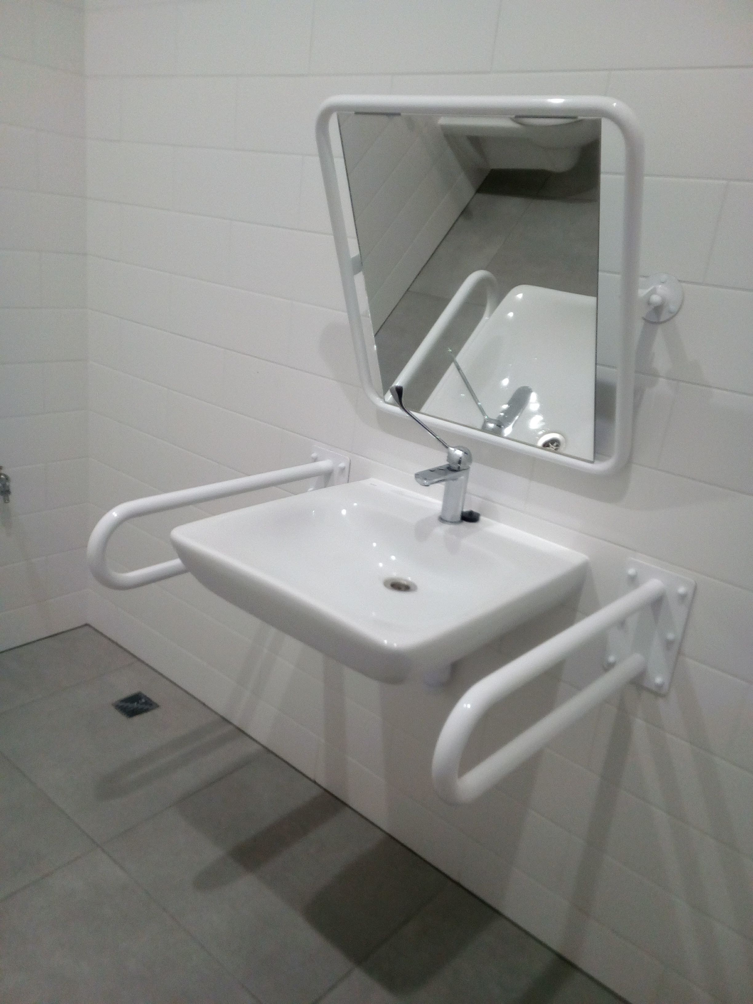Biała umywalka dla niepełnosprawnych z poręczami i lustrem, zamontowana w łazience w Łodzi, widoczna na tle białych płytek.