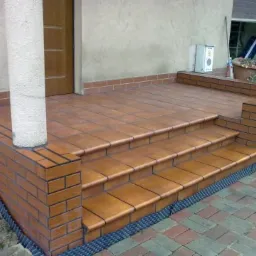 Wykończone schody wejściowe z terakoty i cegły klinkierowej, z widoczną membraną kubełkową przy fundamencie i kolumną podtrzymującą zadaszenie.