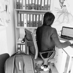 Osoba siedząca przy biurku z laptopem w biurze, za nią regał z segregatorami oznaczonymi nazwami firm i rysunek kobiety w sukni balowej.