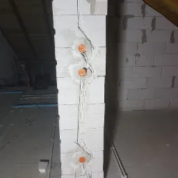 Ściana z betonu komórkowego w trakcie montażu instalacji elektrycznej: widoczne przewody, puszki instalacyjne i fragment drewnianej konstrukcji dachu na poddaszu.