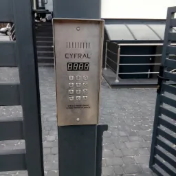 Metalowy panel domofonu Cyfral z klawiaturą numeryczną, zamontowany na grafitowej bramie posesji, z widoczną kostką brukową i fragmentem budynku w tle.