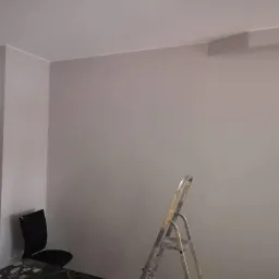 Świeżo pomalowana szara ściana w pokoju, widoczna drabina malarska, czarne krzesło i fragment białej szafy.