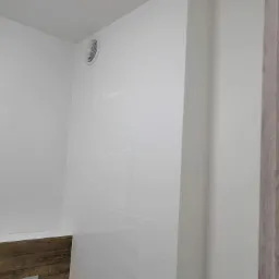 Biała łazienka z drewnopodobnym elementem zabudowy stelaża WC i wentylatorem w suficie, widok z narożnika.