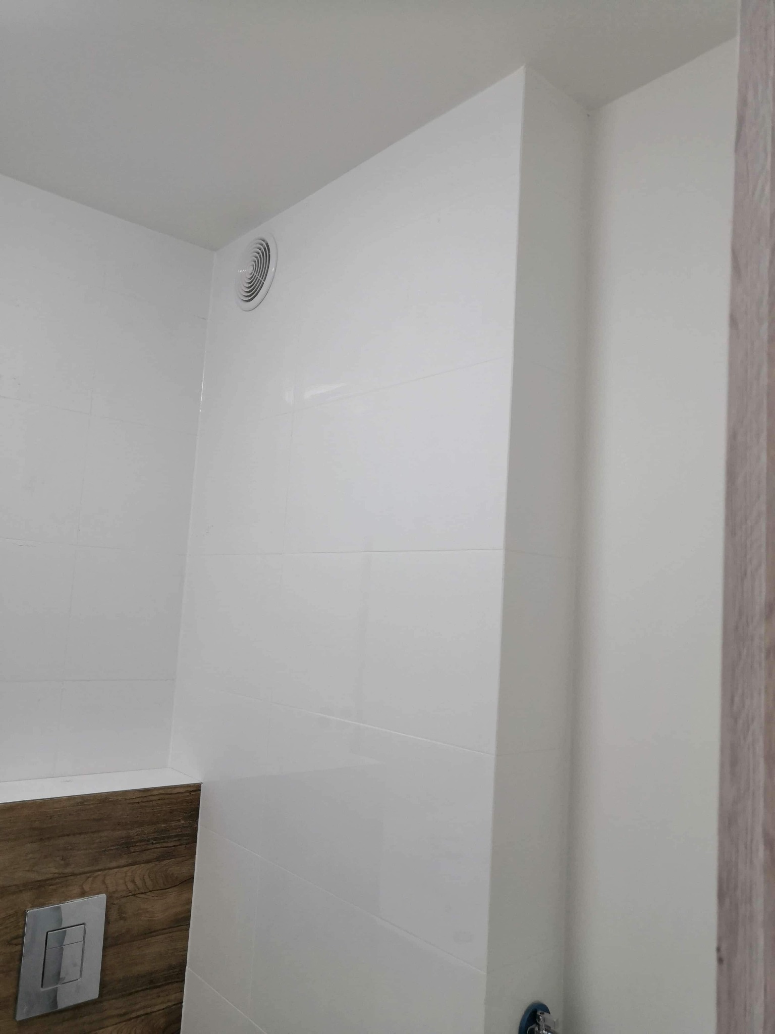 Biała łazienka z drewnopodobnym elementem zabudowy stelaża WC i wentylatorem w suficie, widok z narożnika.