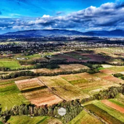 Panoramiczny widok z lotu ptaka na mozaikę pól uprawnych i zabudowań wiejskich, z górami w tle pod dramatycznym, pochmurnym niebem.