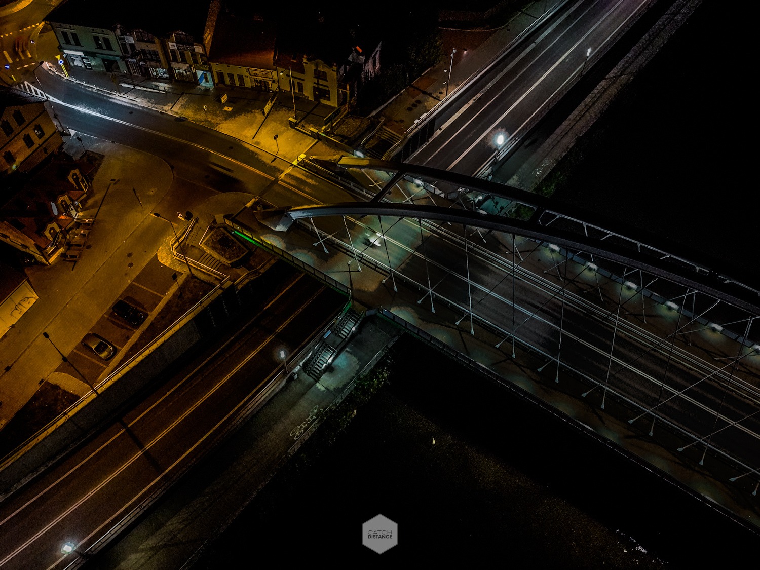 Nocny widok z drona na oświetlony most przecinający rzekę, obok budynki miejskie i ulice z widocznymi smugami świateł samochodowych.