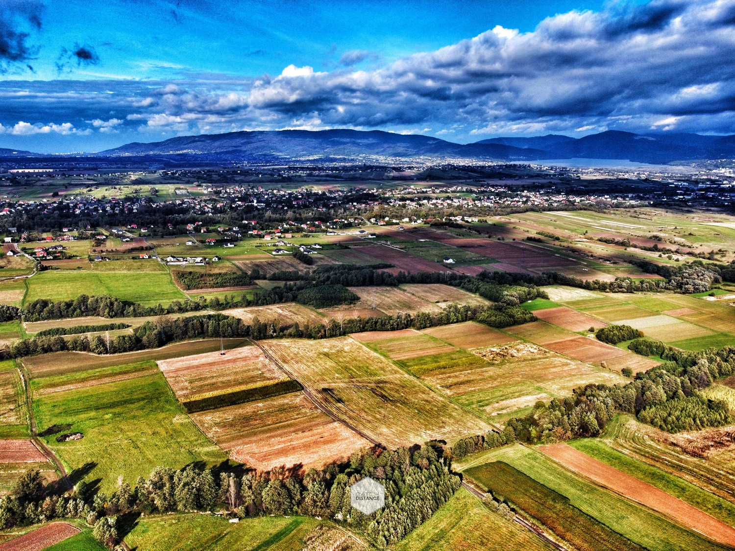 Panoramiczny widok z lotu ptaka na mozaikę pól uprawnych i zabudowań wiejskich, z górami w tle pod dramatycznym, pochmurnym niebem.