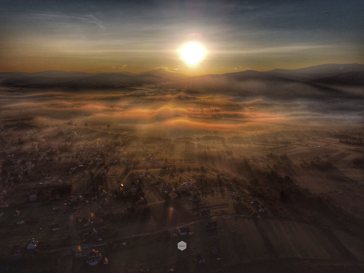 Panoramiczny widok z lotu ptaka na wieś spowitą poranną mgłą, promienie słoneczne przebijają się przez mglisty krajobraz, w tle zarysowane góry na tle wschodzącego słońca.