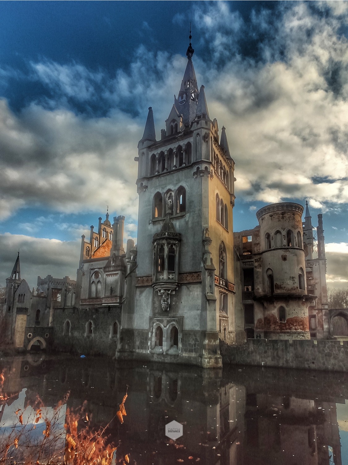 Opuszczony, gotycki zamek odbijający się w wodzie, z widocznymi zniszczeniami i chmurami na niebie.