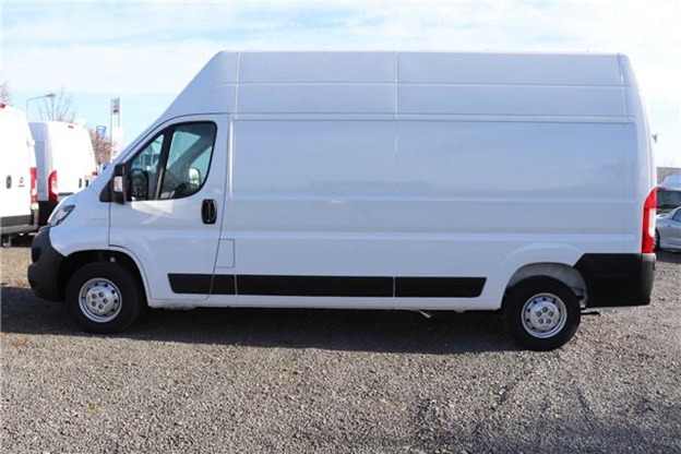 Biały, wysoki furgon dostawczy na parkingu żwirowym, widok z boku, z innymi podobnymi pojazdami w tle.