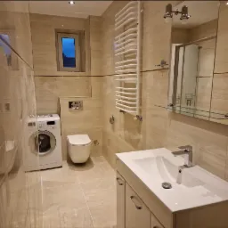 Jasna łazienka po remoncie z beżowymi płytkami, wiszącą toaletą, pralką i grzejnikiem drabinkowym