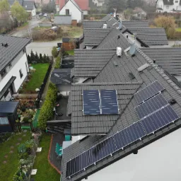 Widok z góry na domy z ciemnymi dachówkami, na jednym z nich panele słoneczne, w tle zabudowa jednorodzinna i zieleń.
