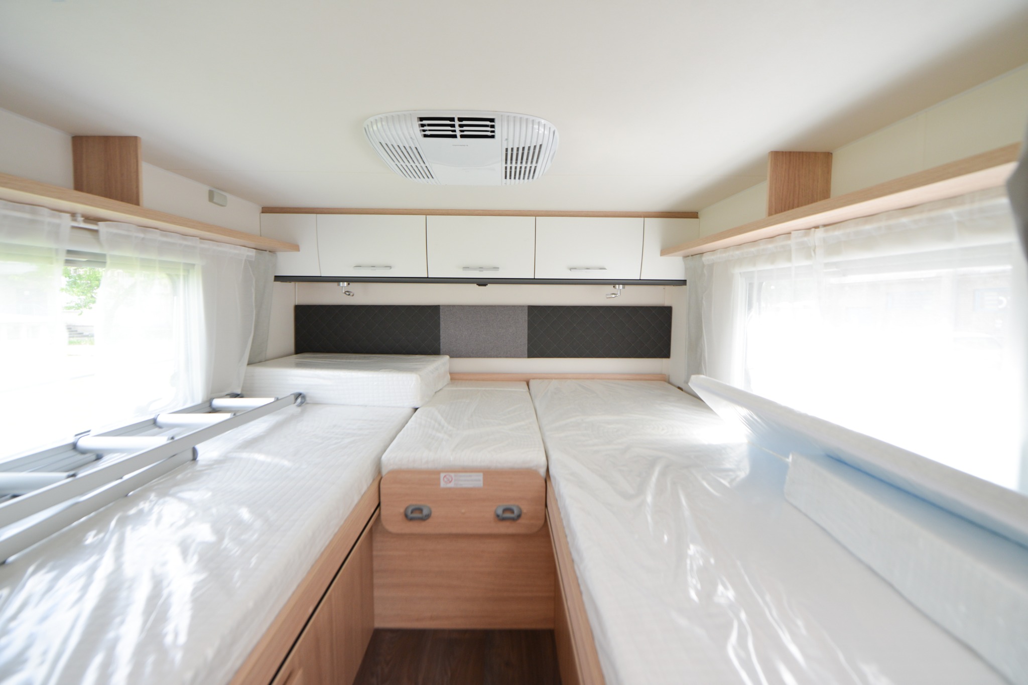 Wnętrze kampera z trzema miejscami do spania, drabinką i klimatyzacją sufitową, wszystko przykryte folią ochronną.