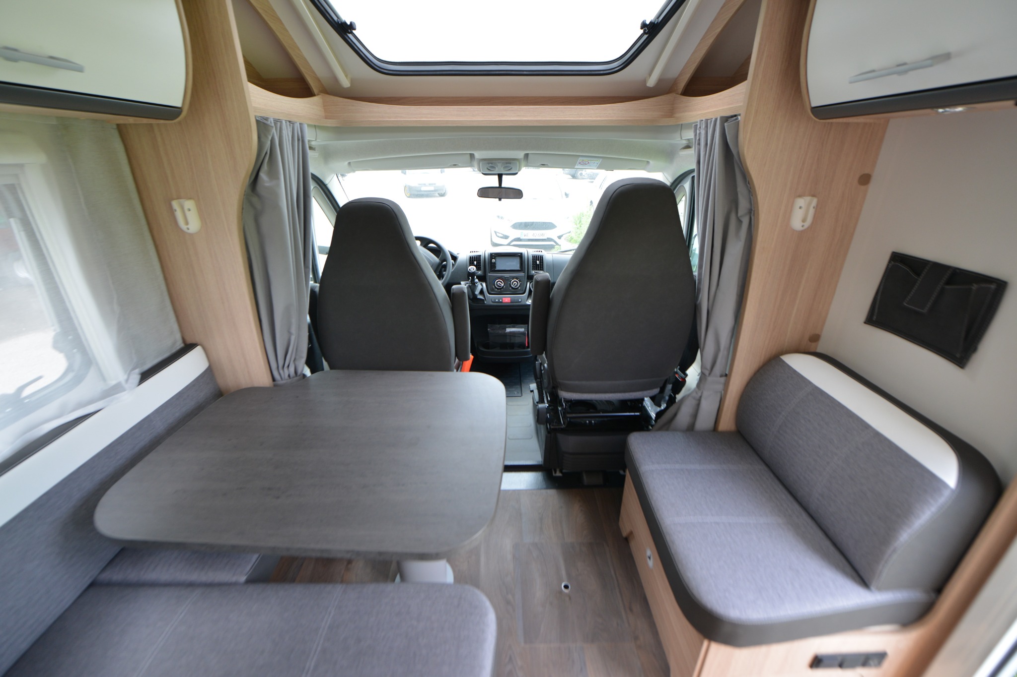 Wnętrze kampera z widokiem na przednie fotele, stolik i kanapy w odcieniach szarości i beżu, oświetlone przez okno dachowe.