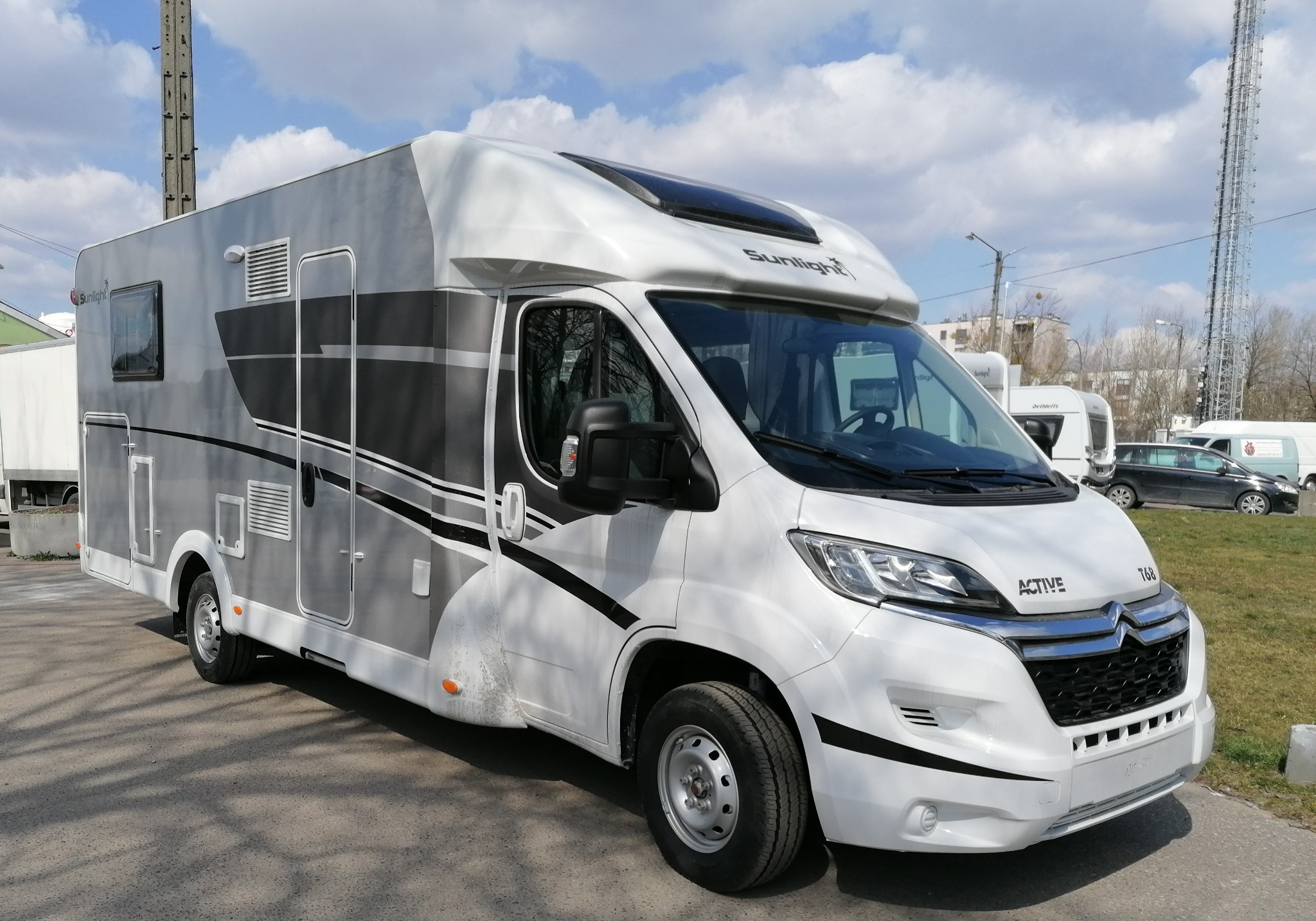 Biało-szary kamper marki Sunlight Active 768 na parkingu, widoczne inne pojazdy i słup wysokiego napięcia w tle.