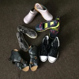 Używane obuwie damskie: para różowych chodaków, espadryle ze wzorem skóry węża, granatowe mokasyny z neonowymi akcentami, czarne trampki i sandały na obcasie z wiązaniami, ułożone na brązowym dywanie.