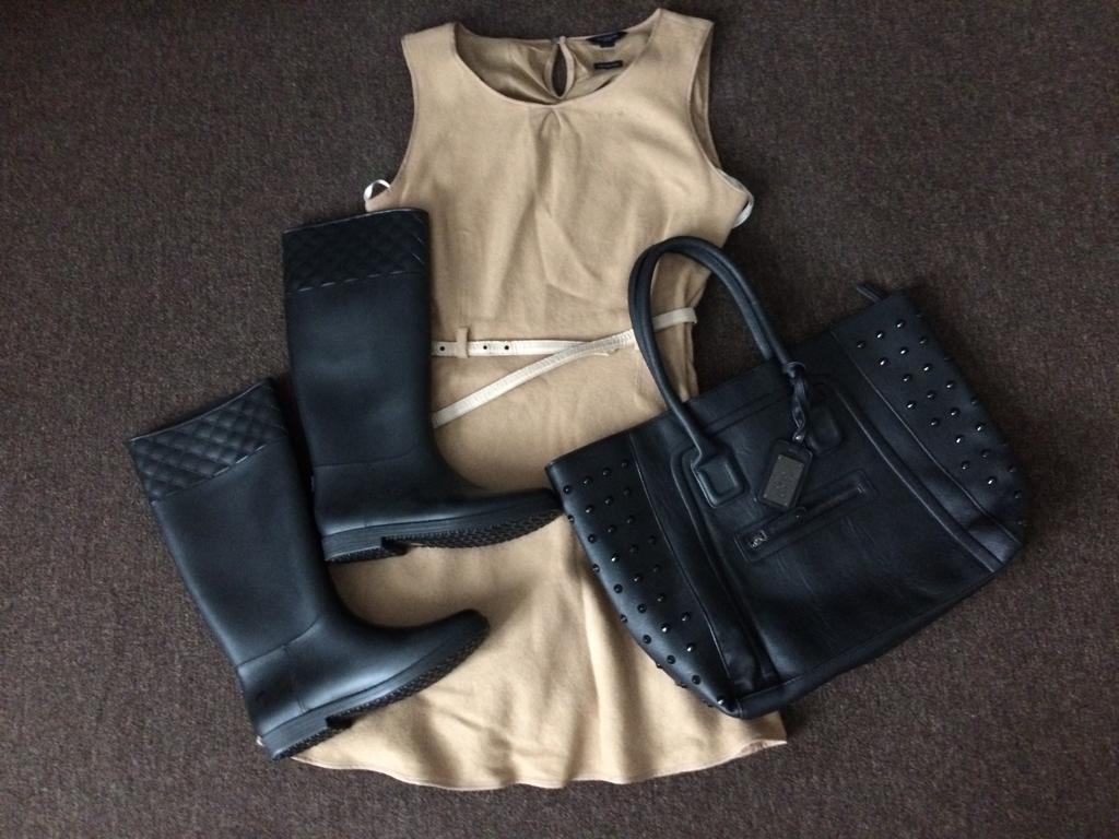 Beżowa sukienka bez rękawów z cienkim paskiem, czarne kalosze z pikowanym detalem oraz czarna torebka z ćwiekami leżące na brązowym dywanie.
