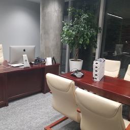 Glassbud - Nowoczesne biuro z drewnianymi biurkami, kremowymi krzesłami, komputerem iMac i dużą rośliną doniczkową przy betonowym filarze i oknie z żaluzjami.