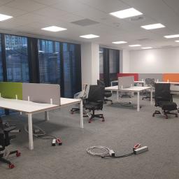 Glassbud - Nowe biuro z białymi biurkami z kolorowymi przegrodami, krzesłami w pokrowcach i widocznymi kablami zasilającymi na szarej wykładzinie dywanowej, widok na przeszklony budynek z żaluzjami.