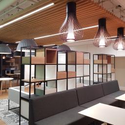 Glassbud - Nowoczesne biuro z drewnianym sufitem, wiszącymi lampami w kształcie klepsydry, regałami z modułowymi półkami i szarą sofą.