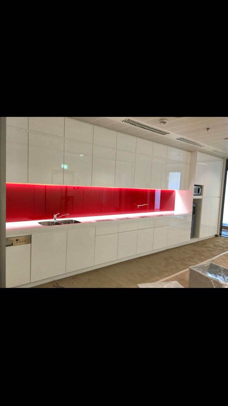 Błyszcząca, biała zabudowa kuchenna w biurze z czerwonym panelem szklanym nad blatem i oświetleniem LED, widoczny zlew i bateria, w tle drzwi i sufit podwieszany.