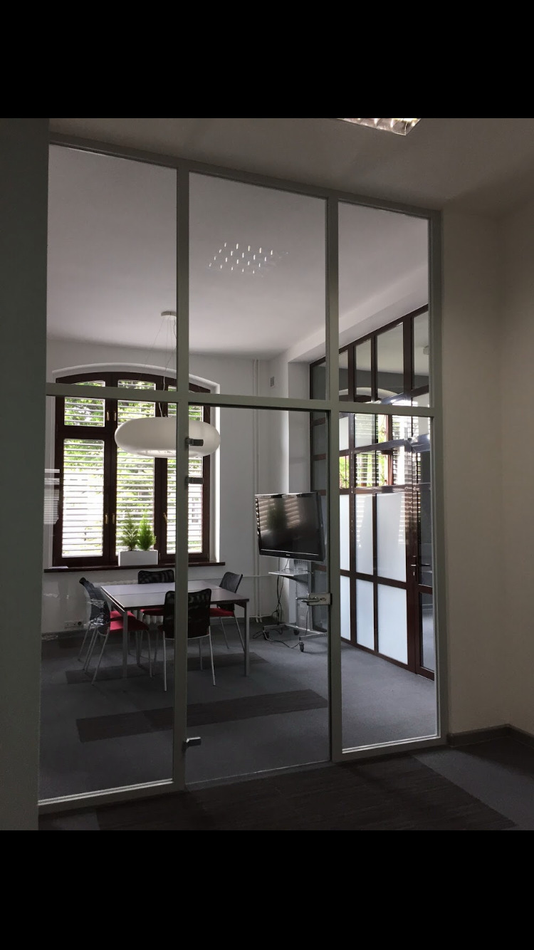 Szklana ściana działowa w biurze; widok na salę konferencyjną z stołem, krzesłami i telewizorem na statywie.