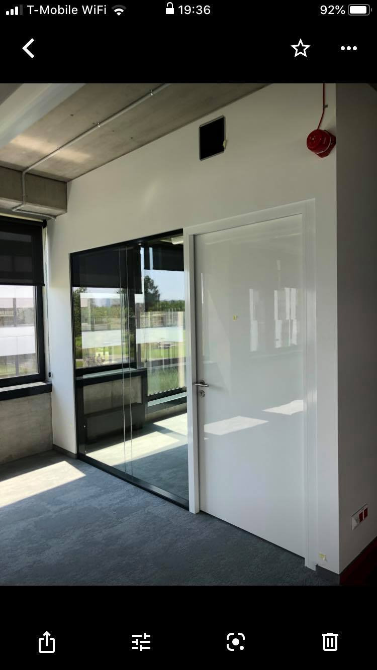 Nowoczesne biuro z przeszkloną ścianą działową i białą, lakierowaną na wysoki połysk, drzwiami, widok na zewnątrz przez okno z roletami.