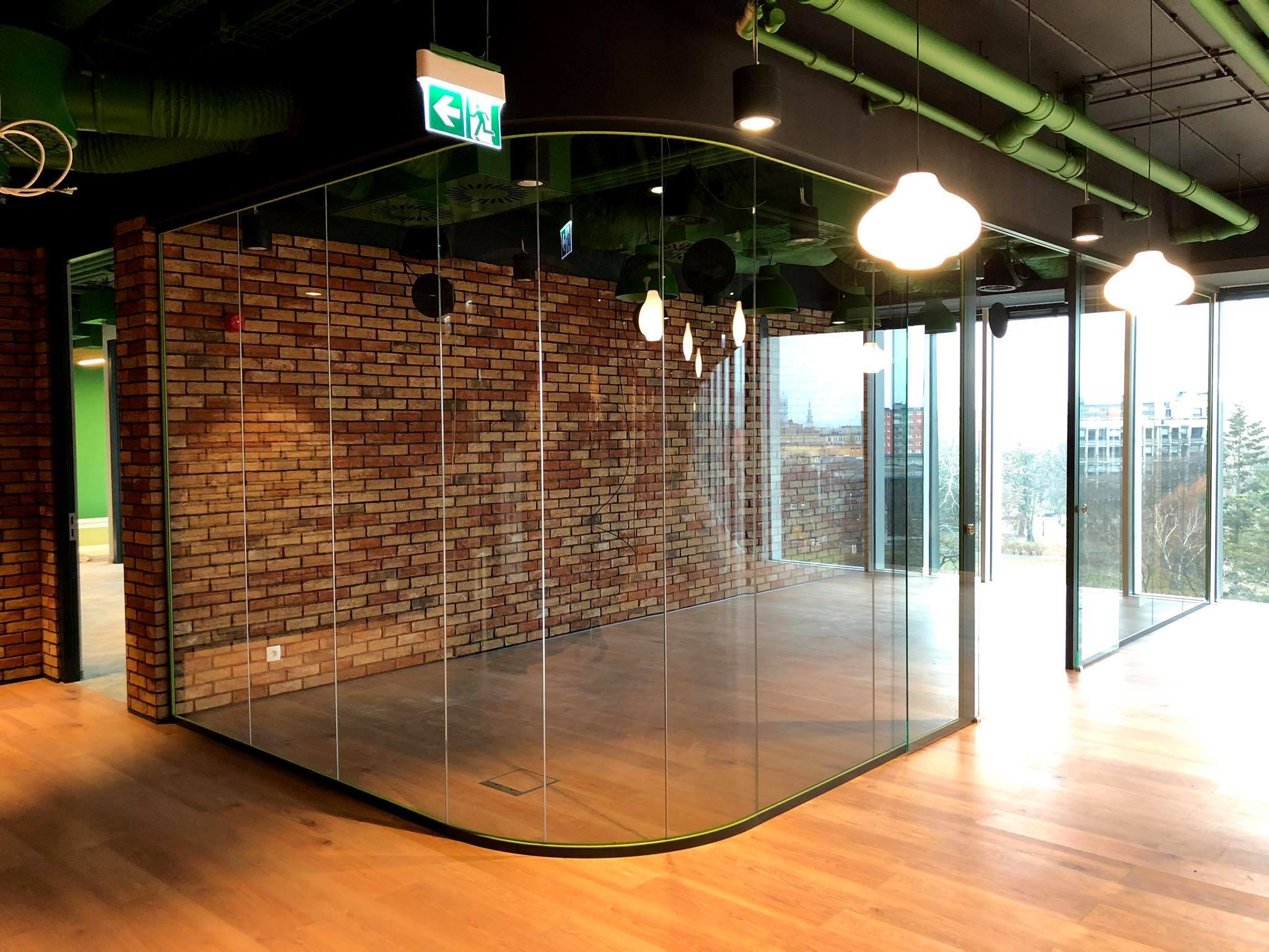 Nowoczesne biuro z przeszkloną ścianą działową, ceglaną ścianą akcentującą i drewnianą podłogą, widoczne elementy instalacji pod sufitem i lampy w kształcie kropli.