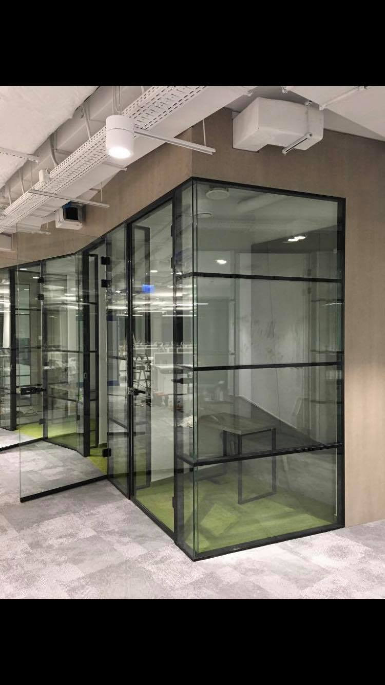Szklane ścianki działowe z czarnymi ramami tworzące przestrzeń biurową z zielonym dywanem i widoczną instalacją sufitową.
