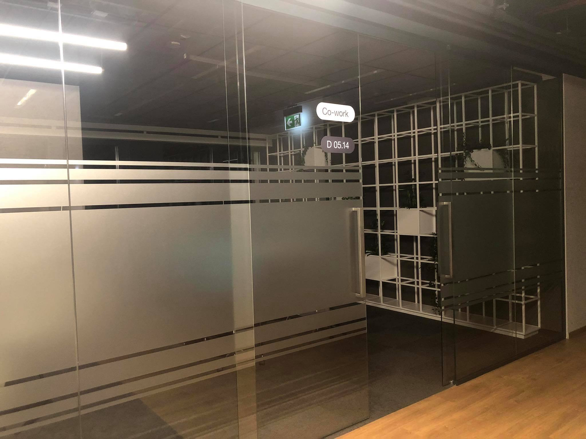 Szklane drzwi do biura typu co-work z poziomymi pasami matowej folii, widoczny znak ewakuacyjny i numer D 05.14, wewnątrz biała konstrukcja regału z roślinami.