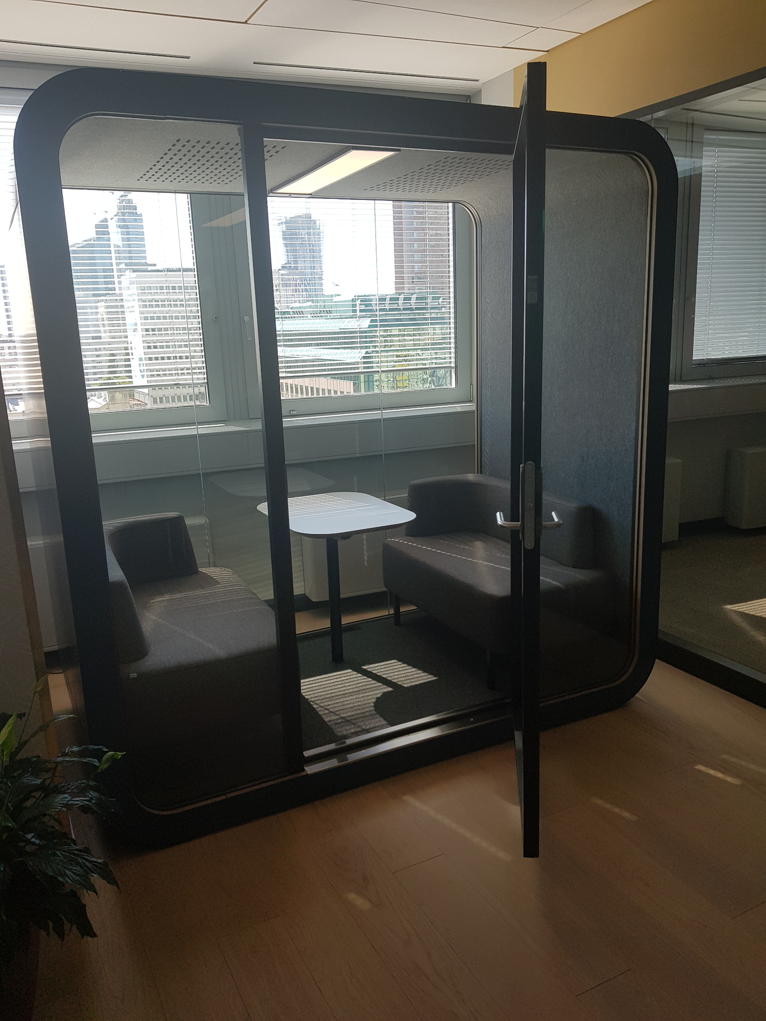 Wyciszona kabina biurowa z otwartymi drzwiami, wewnątrz dwa fotele i mały stolik, widok na miasto przez okno.