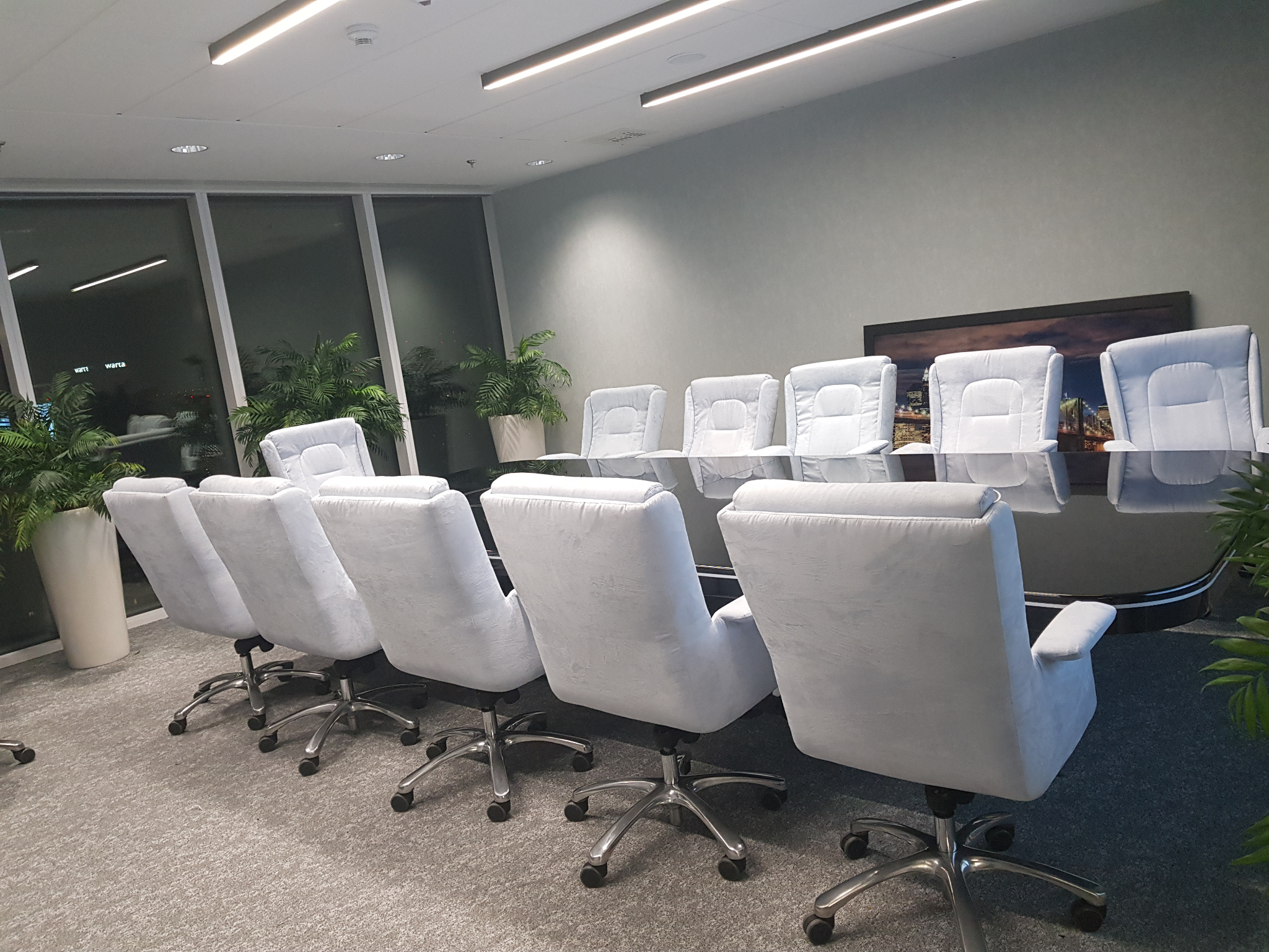 Klimatyczna sala konferencyjna z dużym oknem, roślinami doniczkowymi i szarymi krzesłami obrotowymi wokół czarnego, błyszczącego stołu.