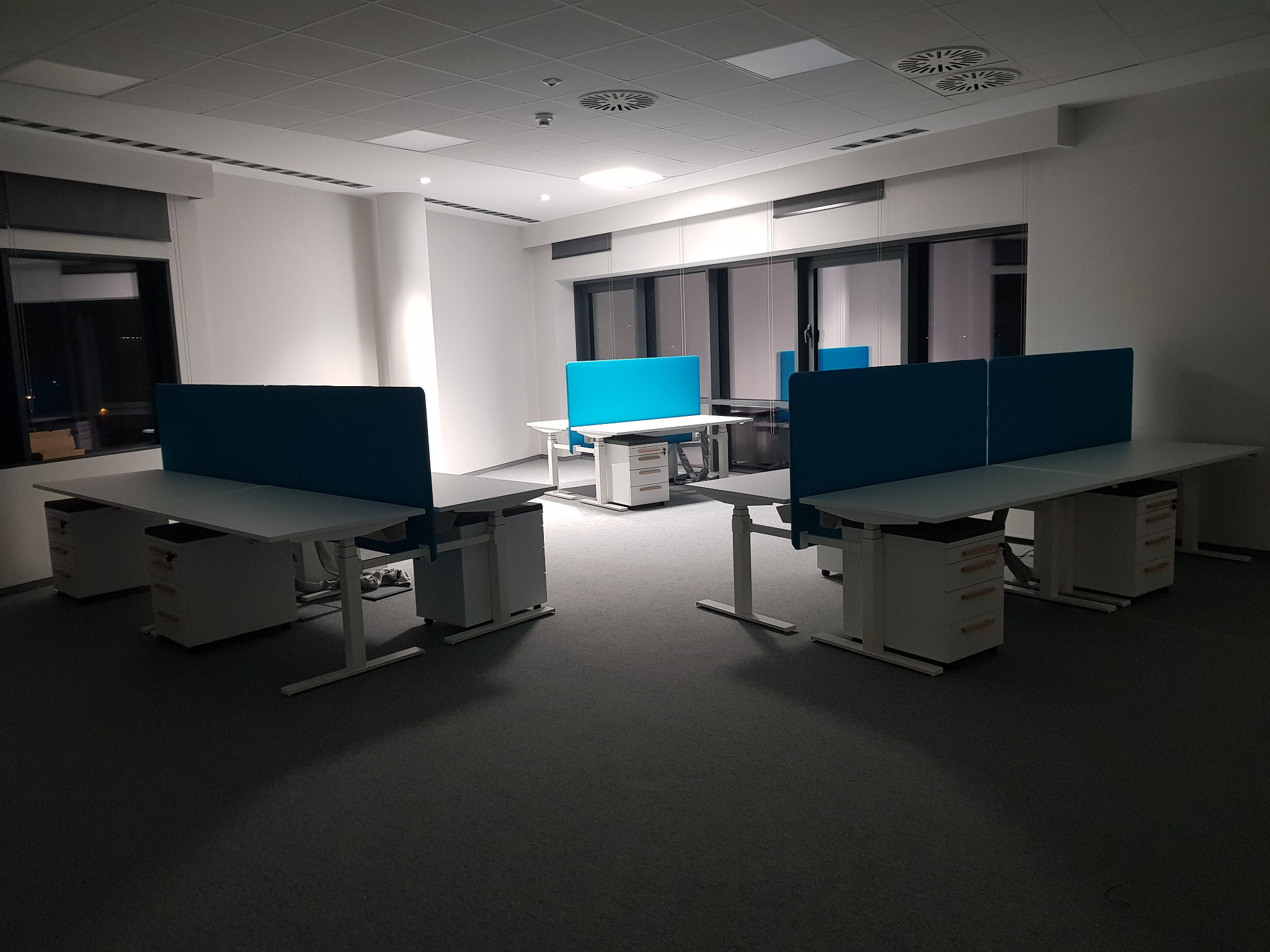 Nowoczesne, puste stanowiska pracy biurowej z białymi biurkami, niebieskimi przegrodami i szarą wykładziną, oświetlone sztucznym światłem.