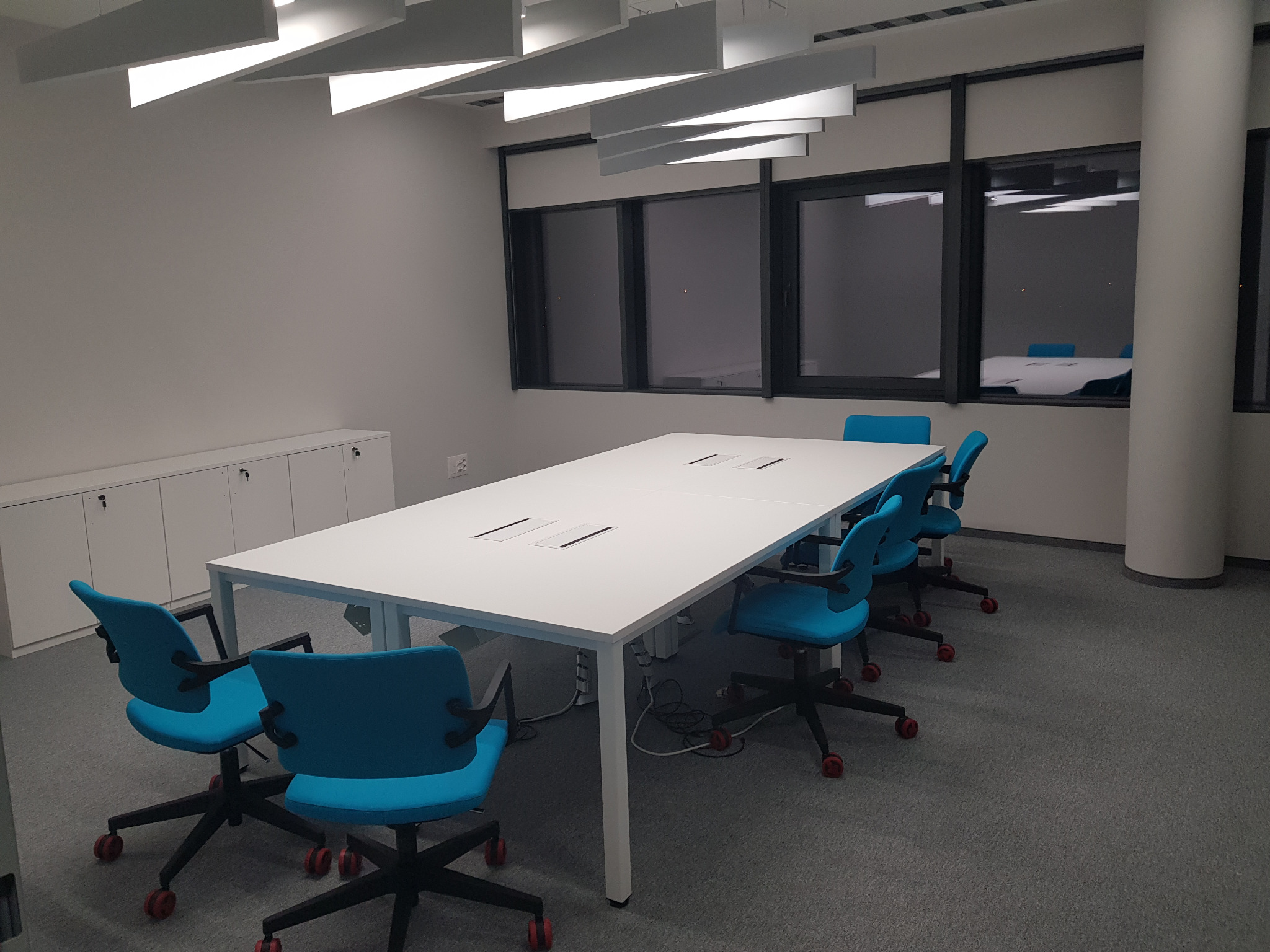 Sala konferencyjna z białym stołem i turkusowymi krzesłami na kółkach z czerwonymi akcentami, oświetlona nowoczesnymi lampami sufitowymi, widok na inne pomieszczenie przez okna.
