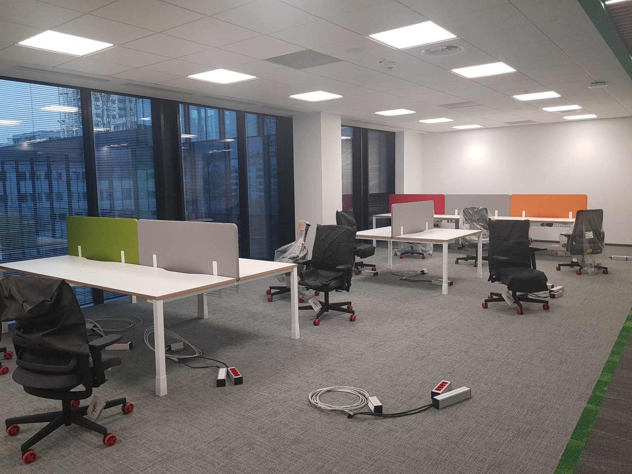 Nowe biuro z białymi biurkami z kolorowymi przegrodami, krzesłami w pokrowcach i widocznymi kablami zasilającymi na szarej wykładzinie dywanowej, widok na przeszklony budynek z żaluzjami.