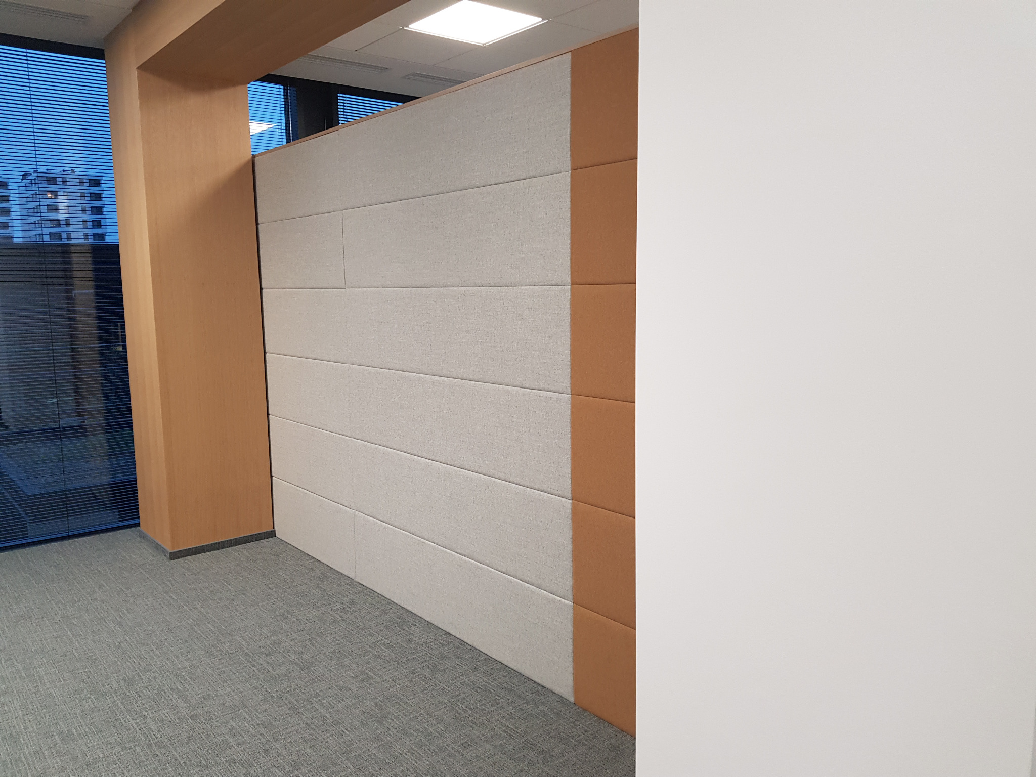 Nowoczesne biuro: panele akustyczne w odcieniach beżu i brązu, dzielące przestrzeń, widok na budynek przez okno z żaluzjami.