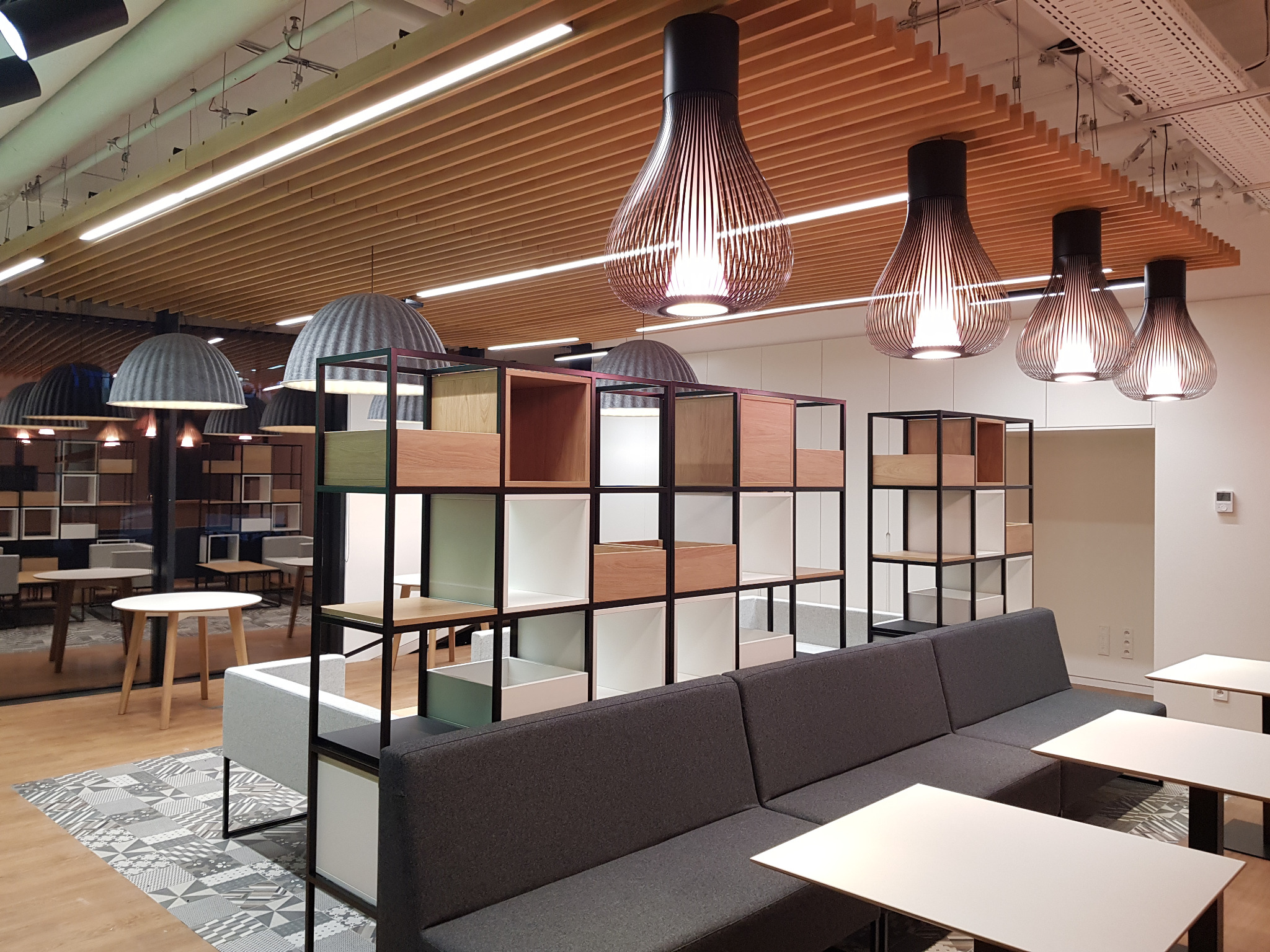 Nowoczesne biuro z drewnianym sufitem, wiszącymi lampami w kształcie klepsydry, regałami z modułowymi półkami i szarą sofą.