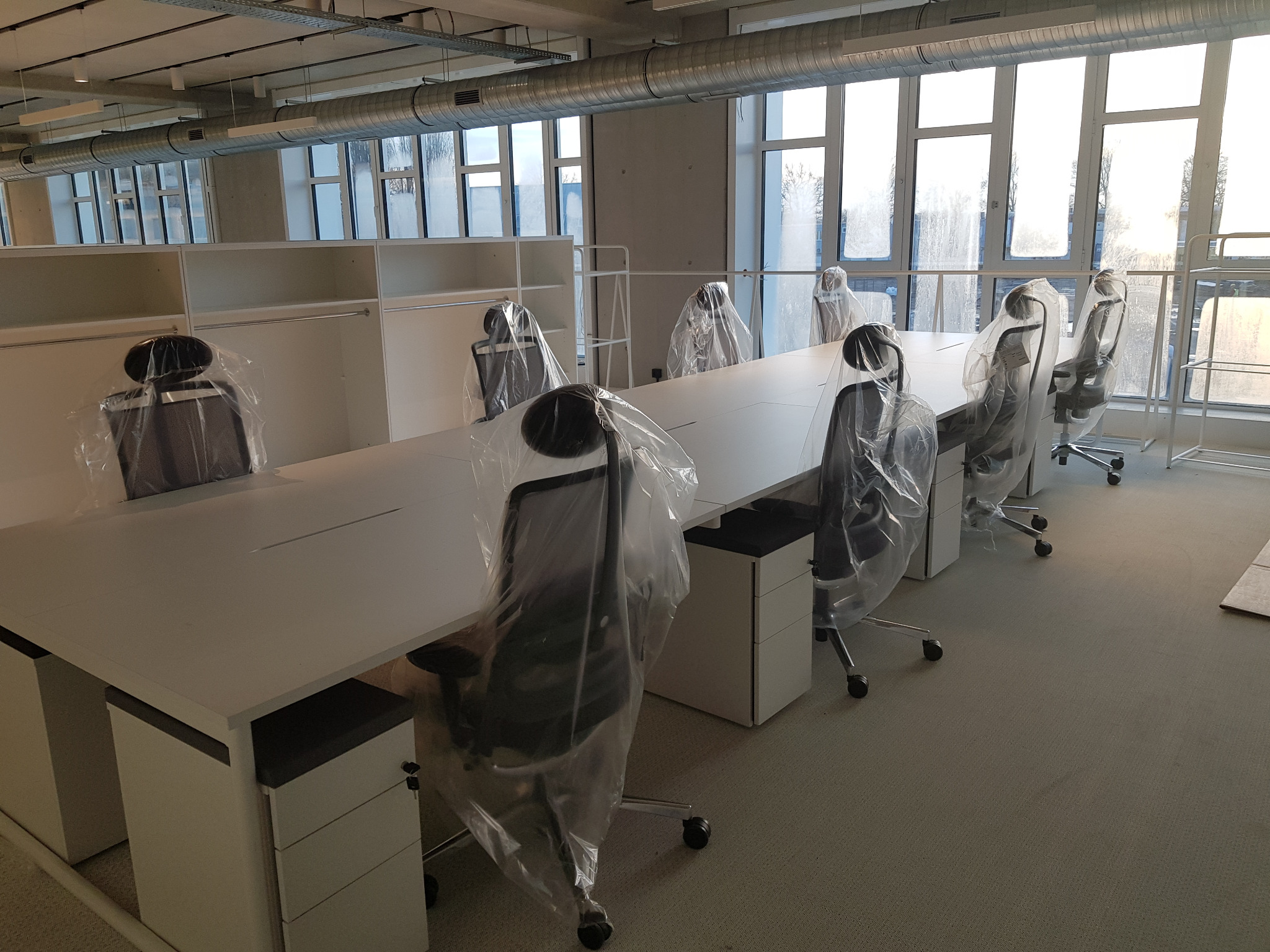 Nowe biuro z długim białym stołem i krzesłami biurowymi w folii ochronnej, przygotowane do użytku, widok na okna i wentylację pod sufitem.