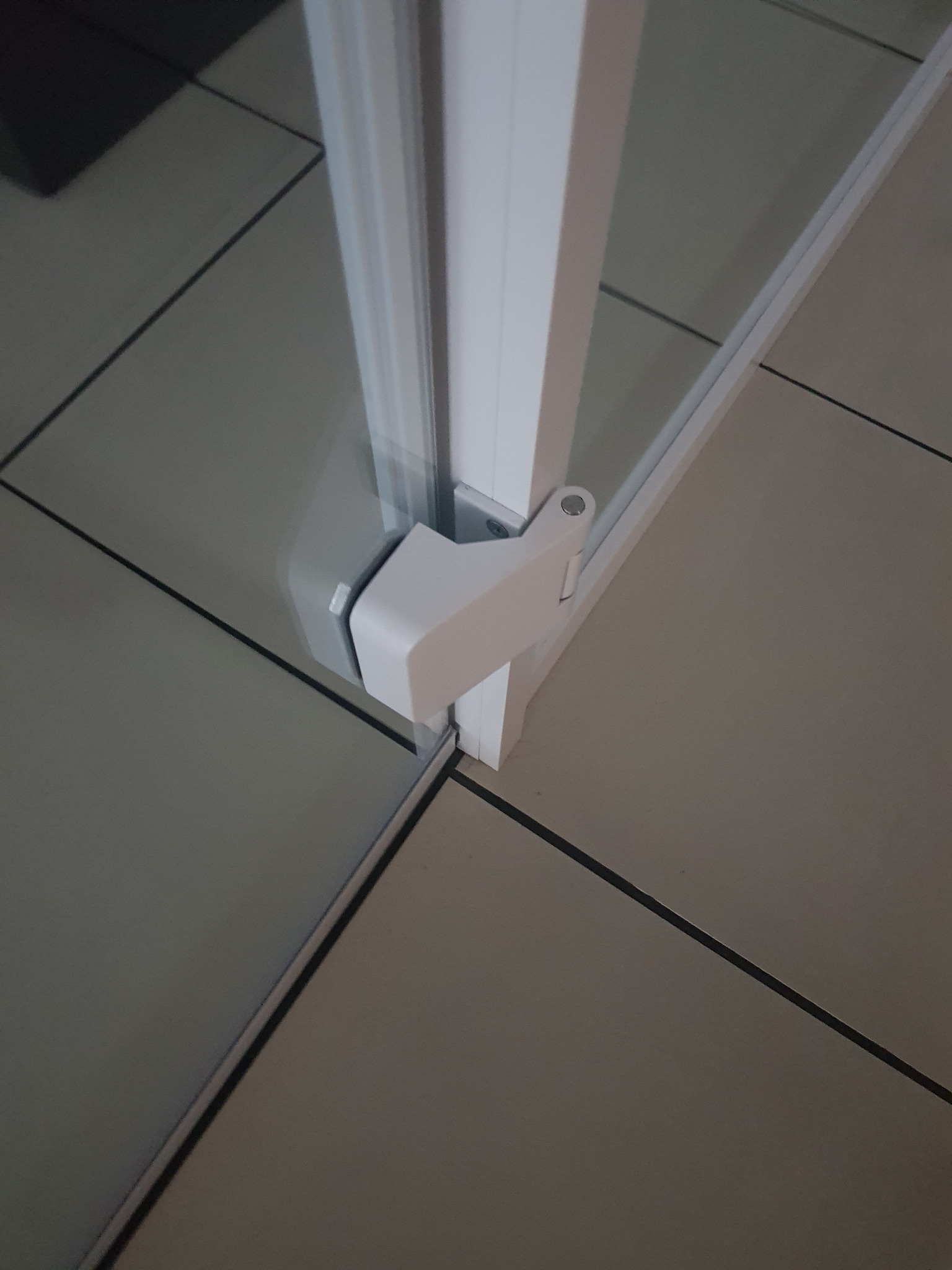 Szczegółowe ujęcie narożnika kabiny prysznicowej z białymi profilami i przezroczystymi szybami na tle płytek podłogowych w odcieniach szarości.