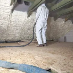 Pracownik w kombinezonie ochronnym aplikuje pianę izolacyjną na poddaszu, widoczne drewniane krokwie i fragment podłogi z płyt OSB.