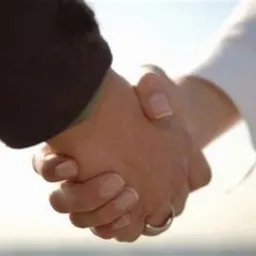 Uścisk dłoni dwóch osób, jedna w ciemnym garniturze, druga w białej koszuli, symbolizujący zawarcie umowy lub partnerstwo, z delikatnym rozmyciem tła.