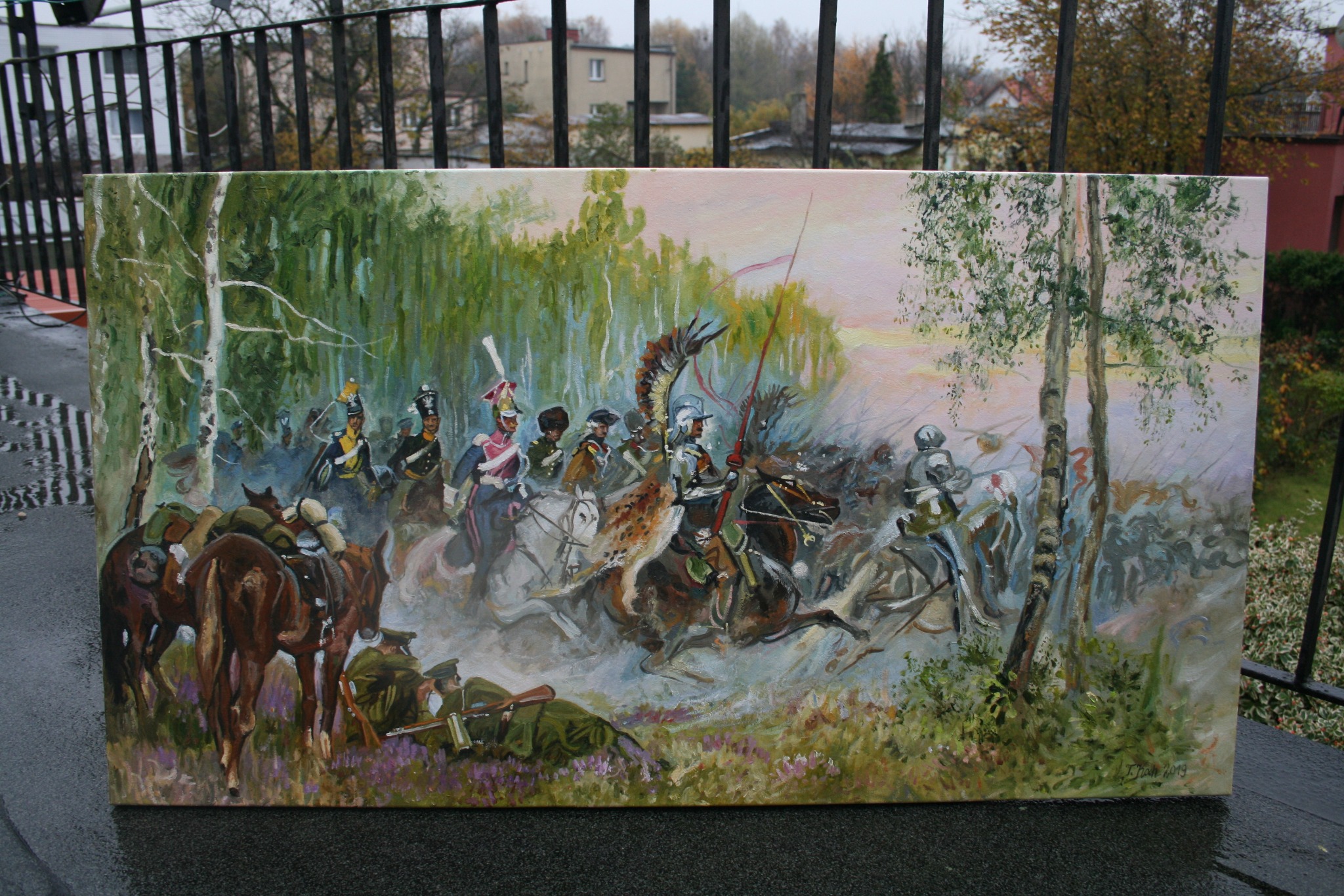 Obraz olejny przedstawiający szarżę husarii na tle lasu, umieszczony na zewnątrz na tle balustrady balkonu i budynków.