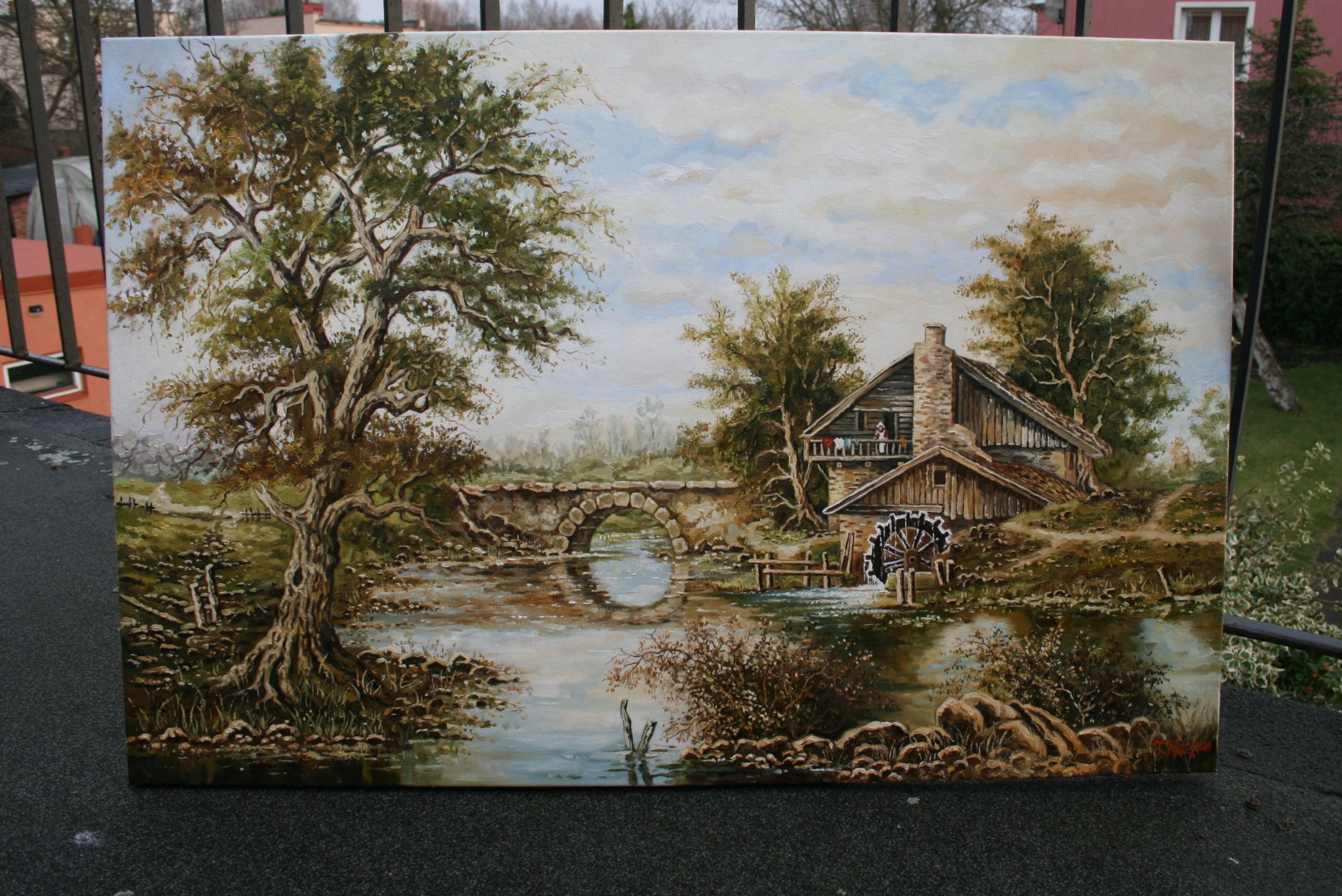 Pejzaż olejny przedstawiający stary młyn wodny z kamiennym mostem łukowym, bujnym drzewem po lewej i osobą na balkonie młyna, umieszczony na tle zewnętrznym.