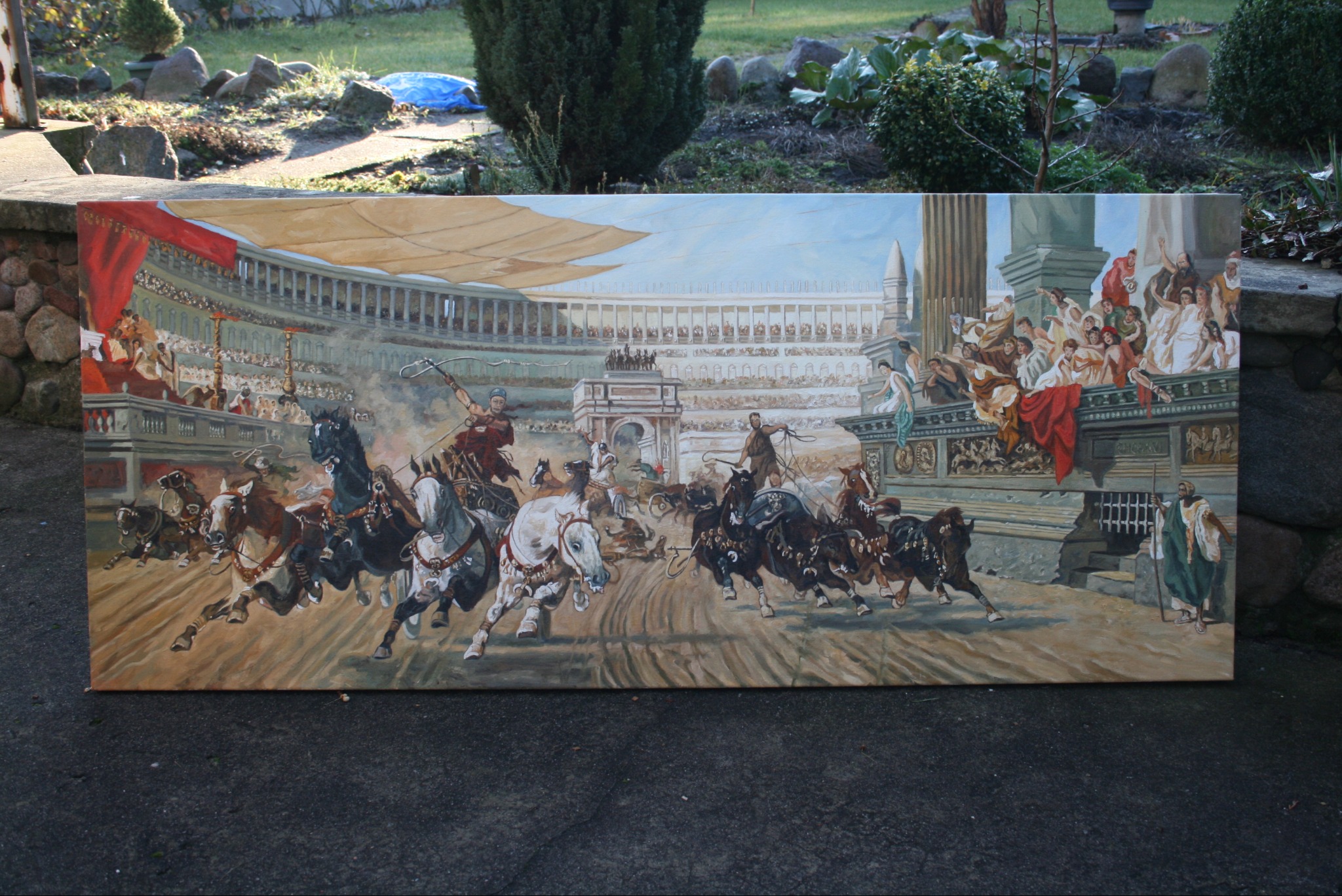 Obraz przedstawia wyścig rydwanów w starożytnym Rzymie, namalowany na dużym płótnie, ustawionym na zewnątrz na szarym podłożu, z widocznym ogrodem w tle.