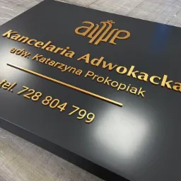 Elegancka tablica informacyjna z nazwą kancelarii adwokackiej, imieniem i nazwiskiem adwokata Katarzyny Prokopiak oraz numerem telefonu, wykonana z czarnego tworzywa z wypukłymi, złotymi literami.