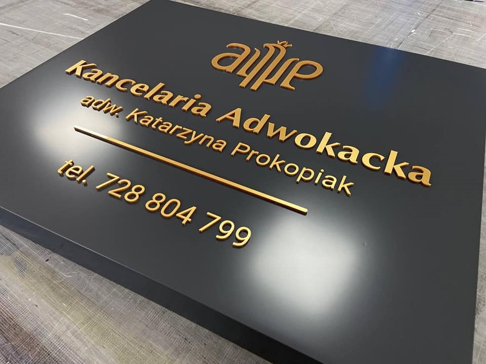 Elegancka tablica informacyjna z nazwą kancelarii adwokackiej, imieniem i nazwiskiem adwokata Katarzyny Prokopiak oraz numerem telefonu, wykonana z czarnego tworzywa z wypukłymi, złotymi literami.