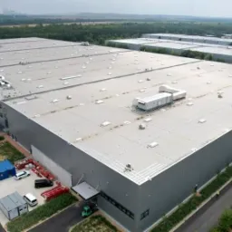 Duży budynek magazynowy z szarej blachy trapezowej, widok z góry, widoczne doki załadunkowe, klimatyzatory na dachu i zaparkowane samochody.