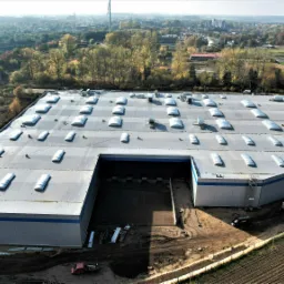 Widok z góry na duży, szary budynek przemysłowy z płaskim dachem pokrytym świetlikami dachowymi; w tle widoczne drzewa i zabudowania miejskie.