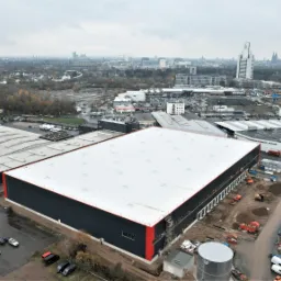Widok z góry na duży budynek magazynowy z białym dachem, na tle panoramy miasta w pochmurny dzień. Wokół budynku zaparkowane samochody i sprzęt budowlany.